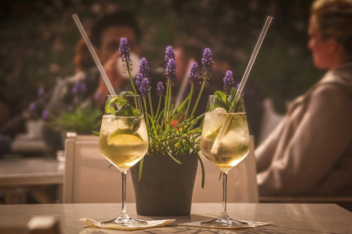 1. Cum se bea prosecco_Tot ce trebuie sa stii despre cocktailuri, combinatii (Hugo, Aperol) si servire corecta_Persoane relaxandu-se la terasa, pahare cu cocktail Hugo Spritz in prim plan, prosecco, lime, menta